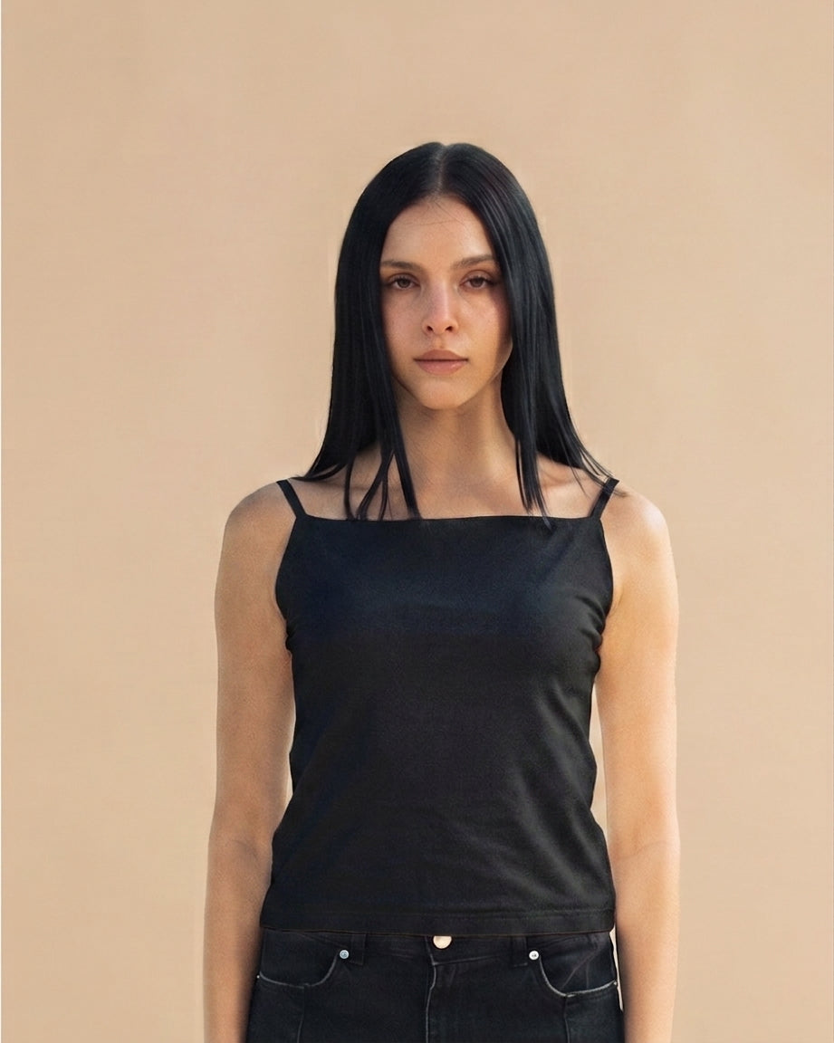 A woman with long black hair wearing a black square-neck camisole top with thin shoulder straps and black jeans, standing against a solid beige background.
