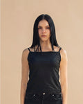 A woman with long black hair wearing a black square-neck camisole top with thin shoulder straps and black jeans, standing against a solid beige background.