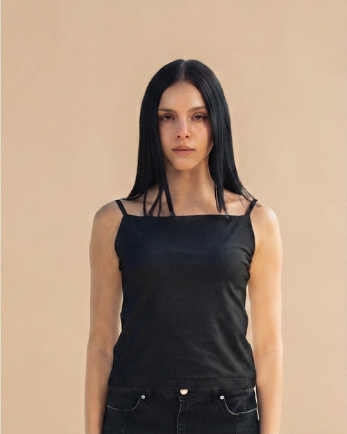 A woman with long black hair wearing a black square-neck camisole top with thin shoulder straps and black jeans, standing against a solid beige background.